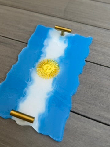 A decorative tray designed like the flag of Argentina, featuring a blue and white wavy border with a gold sun emblem in the center, resting on a gray wooden surface.